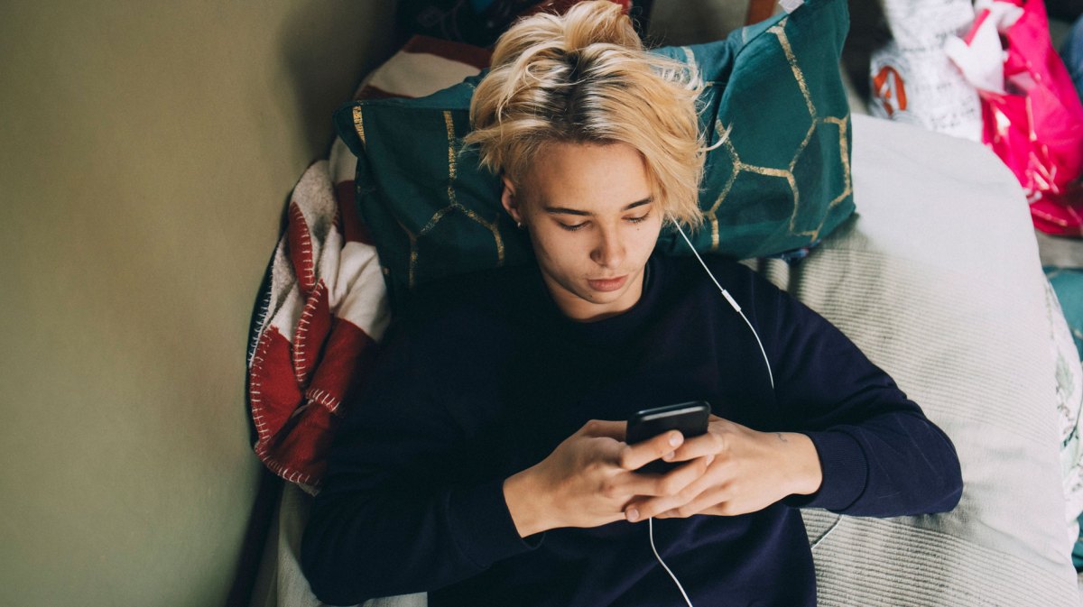 Teenager liegt auf dem Bett, das Handy in der Hand, Kopfhörer im Ohr.
