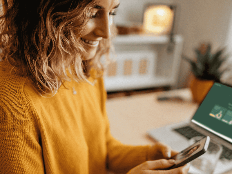 Frau im Homeoffice, trägt gelbes Oberteil und hält Smartphone in der Hand