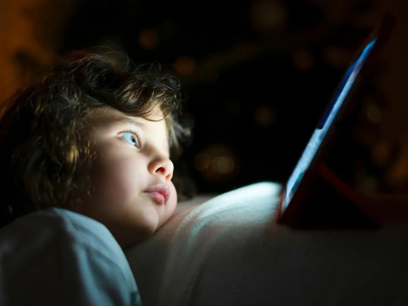 Kind, ca. 4 Jahre, sitzt im Dunkeln gebannt und ganz nah vor einem Tablet-Bildschirm.