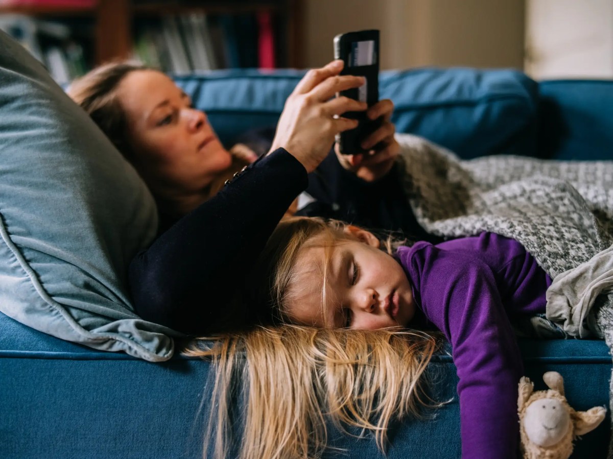Mutter liegt auf der Couch und benutzt ihr Handy. Tochter liegt daneben und schläft