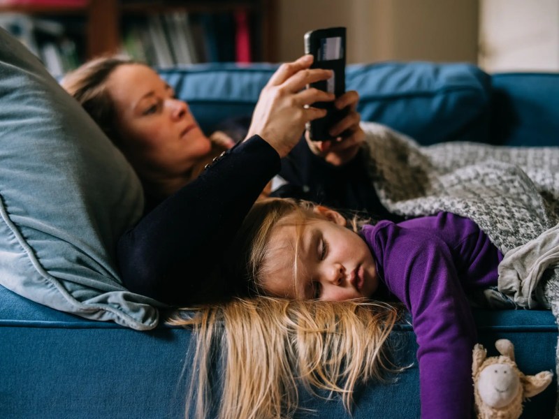Mutter liegt auf der Couch und benutzt ihr Handy. Tochter liegt daneben und schläft