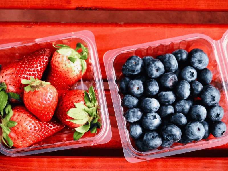 Geöffnete Plastikschalen mit Erdbeeren und Blaubeeren auf rotem Holztisch.