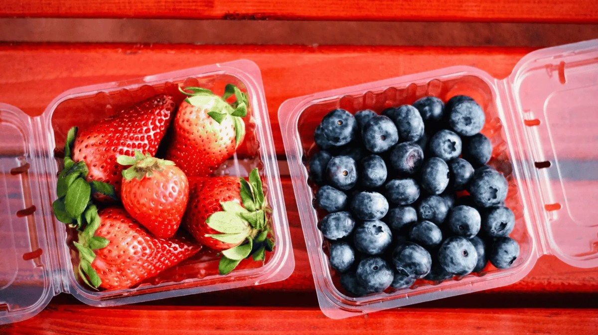 Geöffnete Plastikschalen mit Erdbeeren und Blaubeeren auf rotem Holztisch.