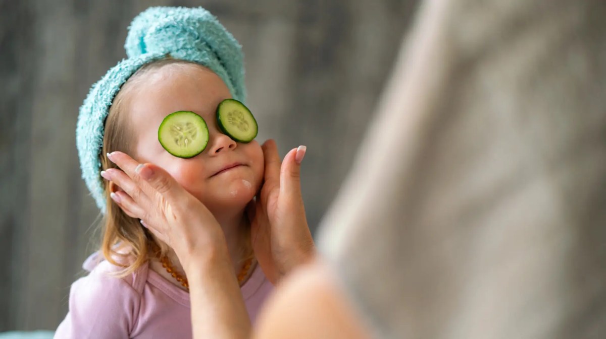 Kind mit Handtuch im Haar und Gurkenscheiben auf den Augen wird im Gesicht eingecremt.