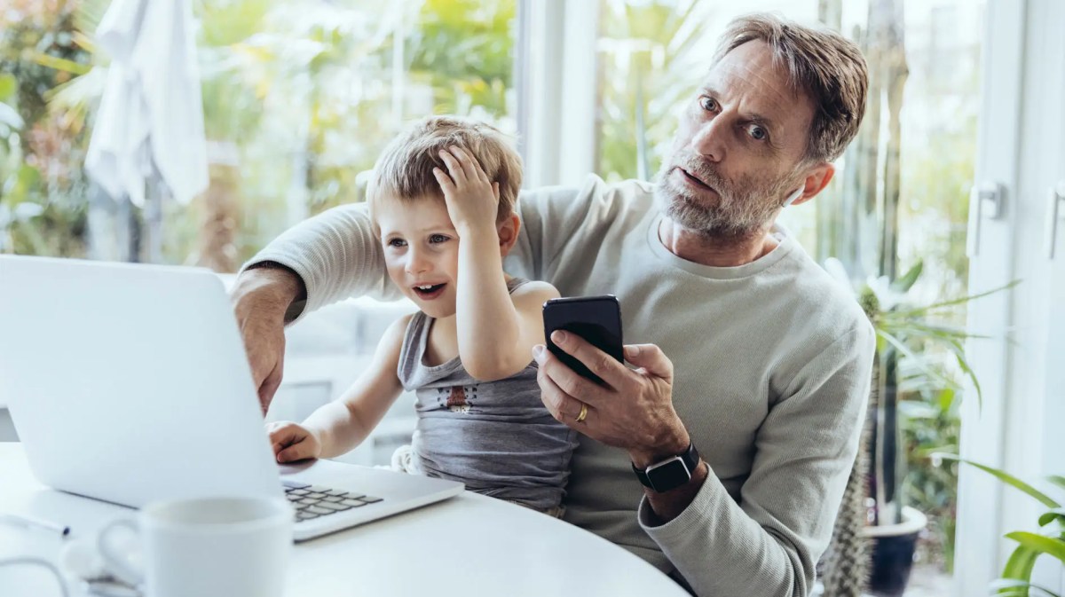 Vater mit Handy in der Hand schaut erschrocken und hat seinen Sohn auf dem Schoß, der etwas am Laptop macht.