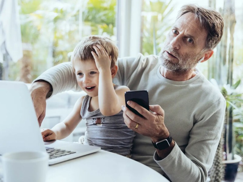 Vater mit Handy in der Hand schaut erschrocken und hat seinen Sohn auf dem Schoß, der etwas am Laptop macht.