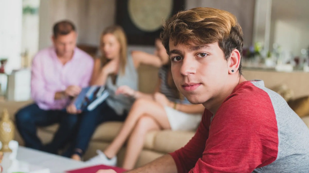 Teenagersohn sitzt im Vordergrund und schaut ernst. Eltern und Schwester sitzen verschwommen im Hintergrund.