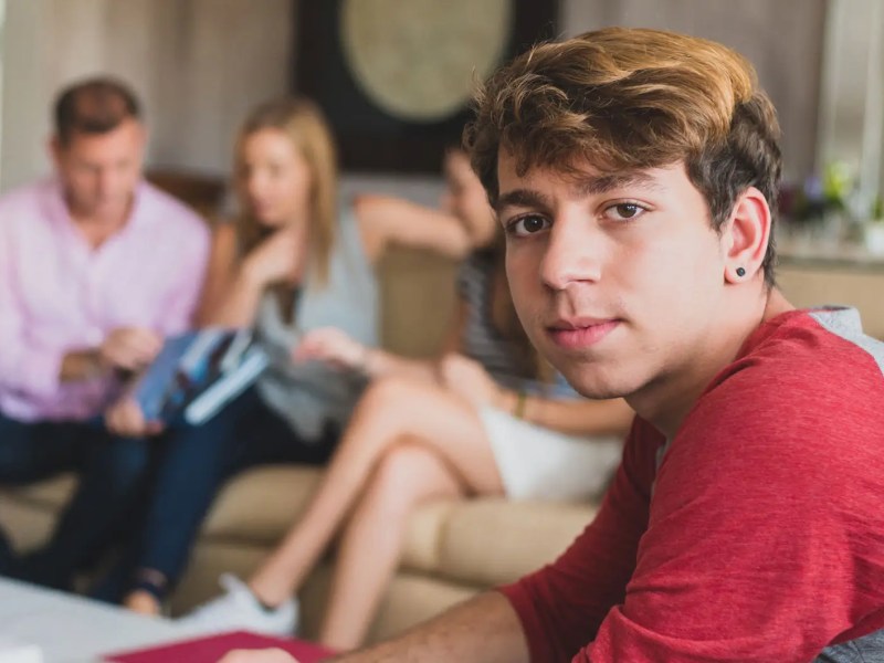 Teenagersohn sitzt im Vordergrund und schaut ernst. Eltern und Schwester sitzen verschwommen im Hintergrund.