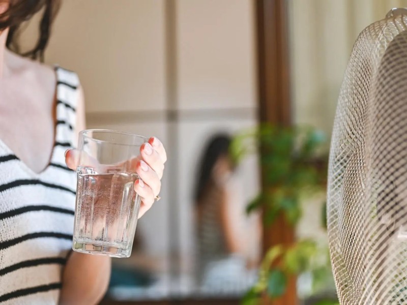 Frau mit einem Glas Wasser kühlt sich vor dem Ventilator ab.