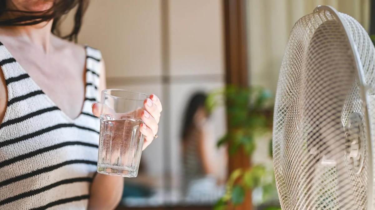Frau mit einem Glas Wasser kühlt sich vor dem Ventilator ab.