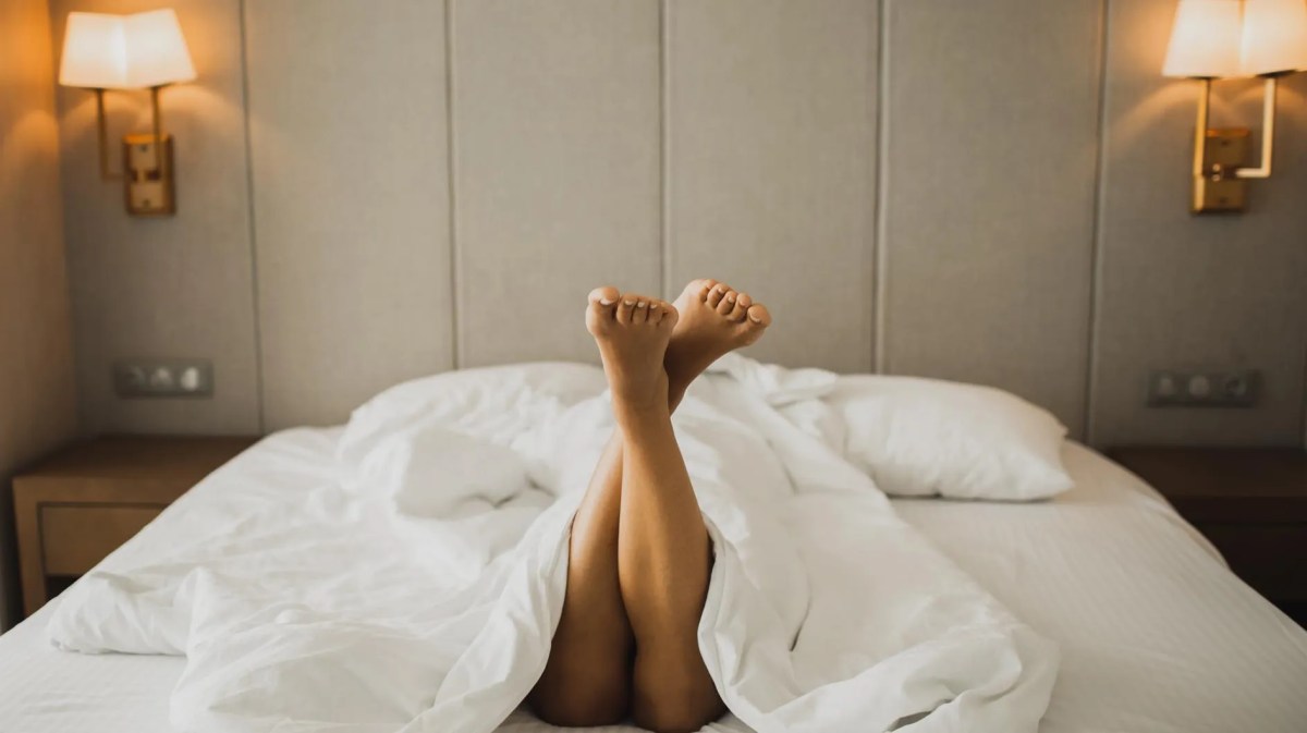 Frau liegt in einem Hotelbett, nackte Beine gucken unter der Decke hervor
