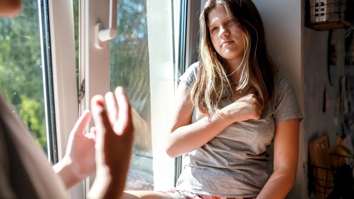 Mädchen sitzt genervt auf der Fensterbank, ihr gegenüber ist eine Person angeschnitten zu erkennen.
