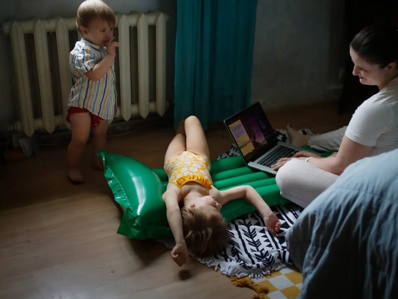 Mutter arbeitet am Laptop, w&auml;hrend ihre Kinder um sie herum spielen.