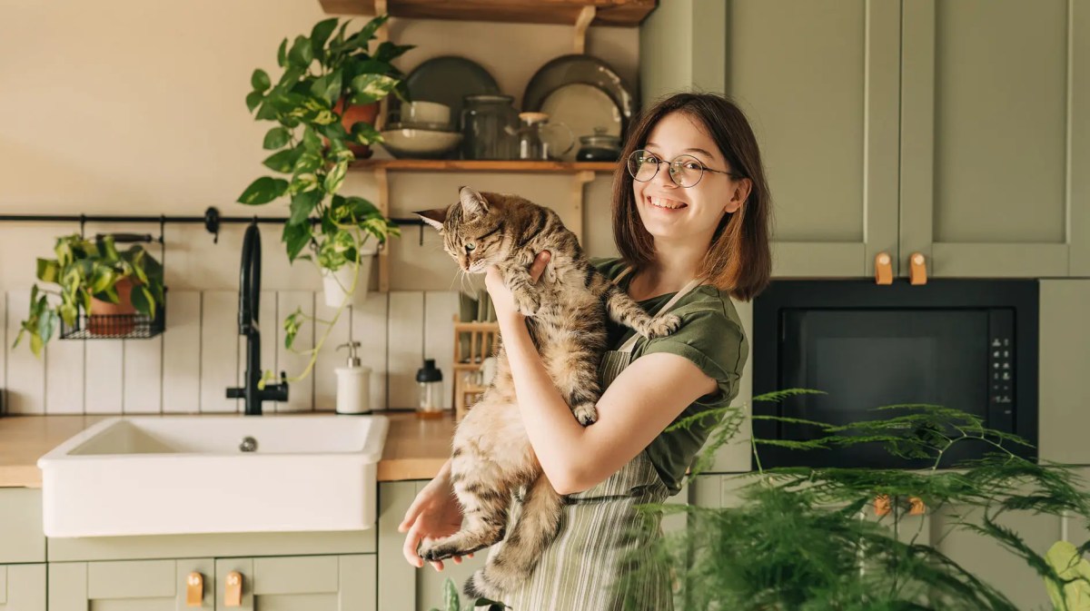Teenagermädchen steht lachend in der Küche und hält eine Katze auf dem arm.