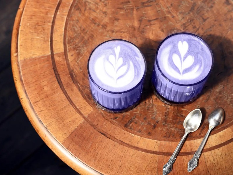 Zwei Ube Latte in violetter Farbe auf einem Holztisch mit zwei silbernen L&ouml;ffeln.