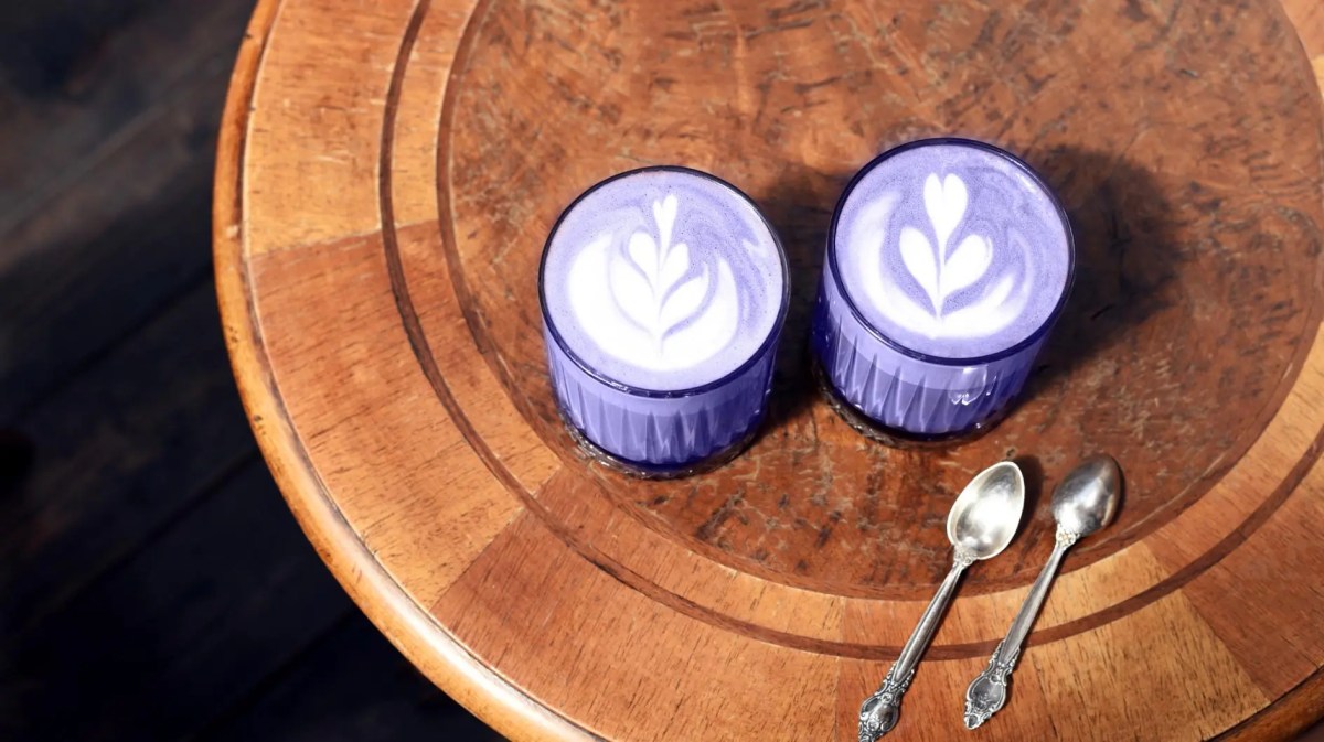 Zwei Ube Latte in violetter Farbe auf einem Holztisch mit zwei silbernen Löffeln.