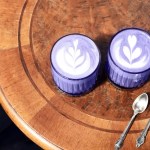 Zwei Ube Latte in violetter Farbe auf einem Holztisch mit zwei silbernen Löffeln.