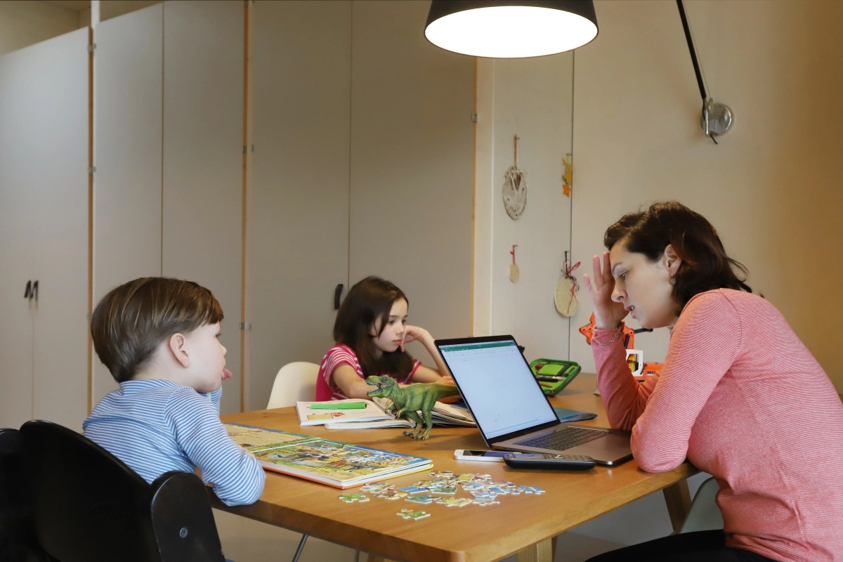 Mutter sitzt gestresst vor einem Laptop zusmamen mit ihren zwei Kindern, die spielen.