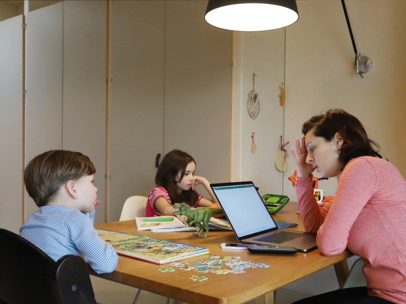 Mutter sitzt gestresst vor einem Laptop zusmamen mit ihren zwei Kindern, die spielen.