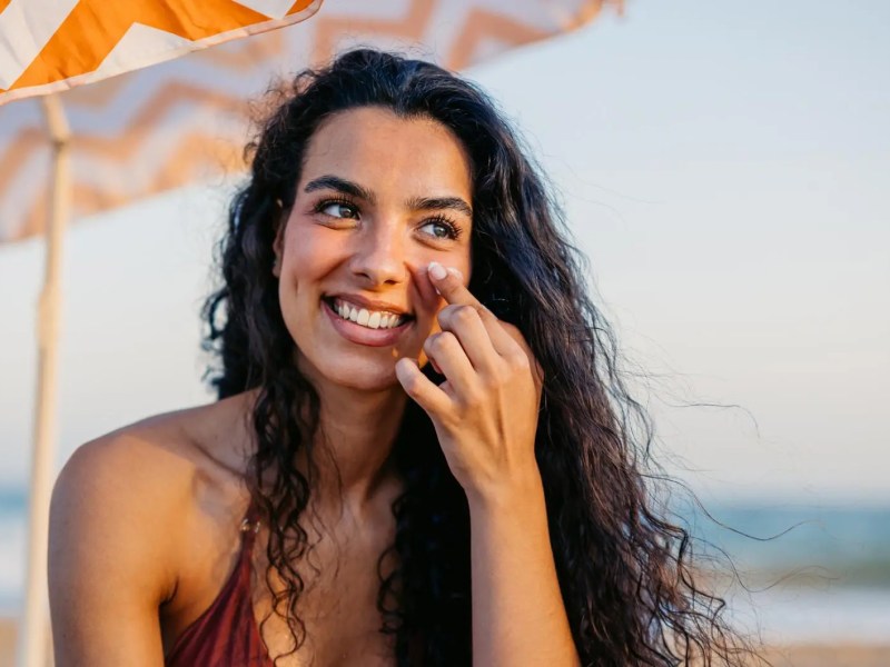 Junge Frau mit braunen Haaren sitzt am Strand und tr&auml;gt Sonnencreme auf.