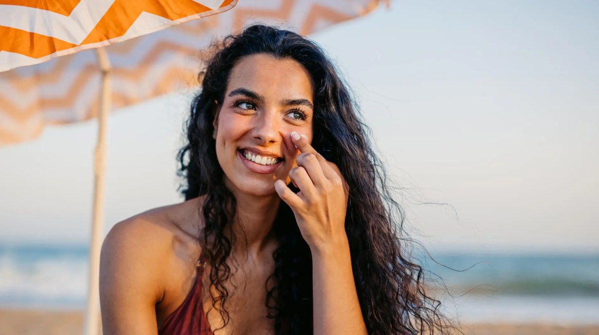 Junge Frau mit braunen Haaren sitzt am Strand und trägt Sonnencreme auf.