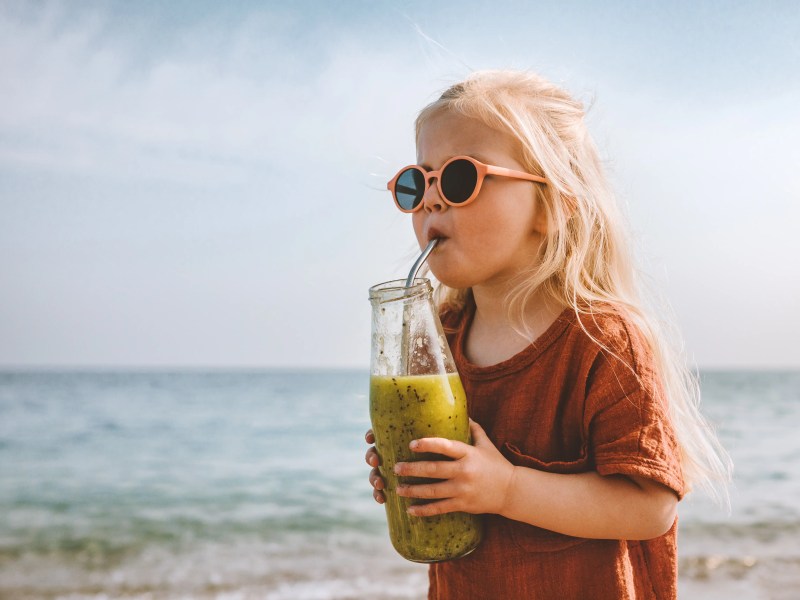 Klienes Mädchen mit Sonnebrille steht am Strand mit einem Glas in der Hand un trinkt aus einem Strohhalm