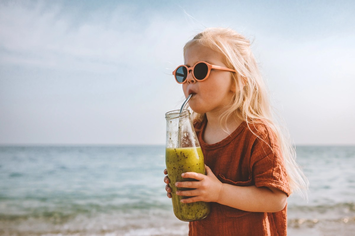 Klienes Mädchen mit Sonnebrille steht am Strand mit einem Glas in der Hand un trinkt aus einem Strohhalm