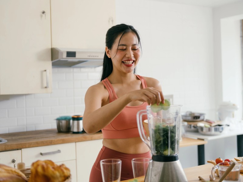 Junge Frau steht in der Küch eund pürriert sich einen gesunden, grünen Smoothie mit Gemüse.