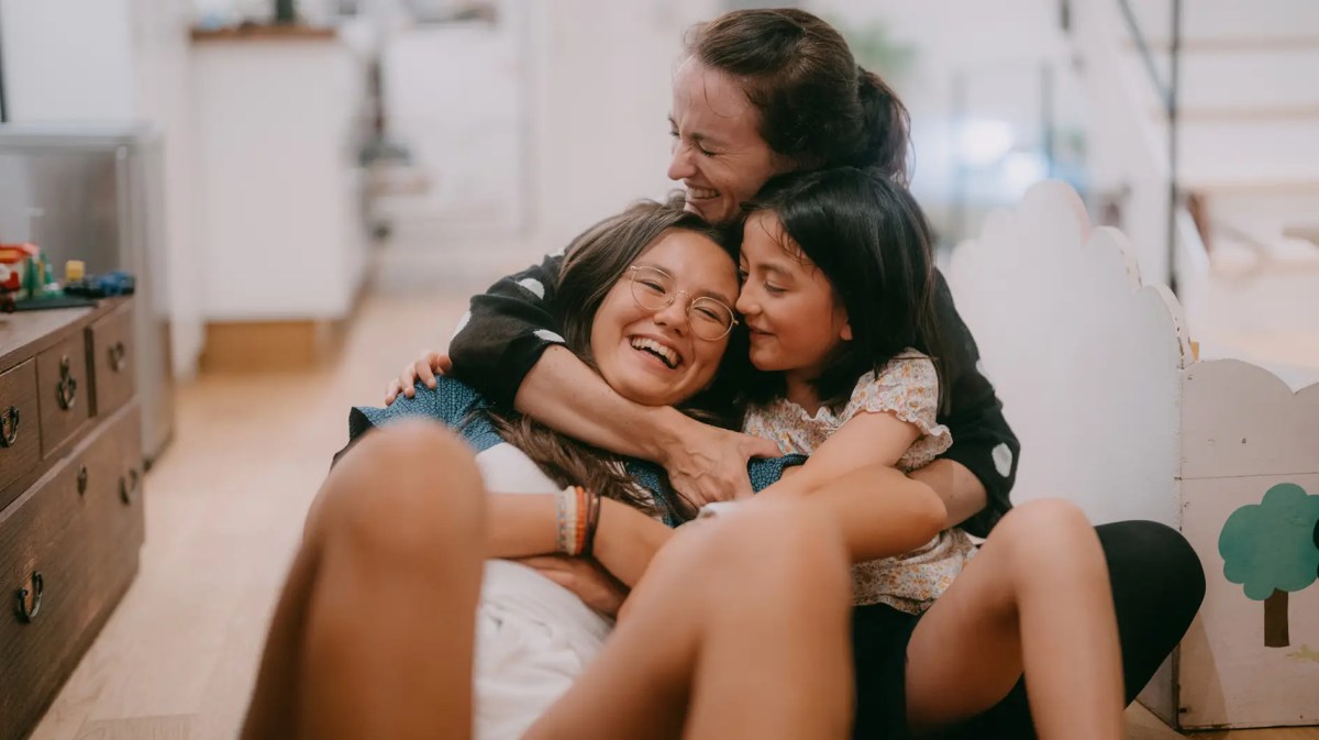 Mutter hält ihre Teenagertochter und eine jüngere Tochter im Arm, alle sitzen auf dem Boden und lachen.