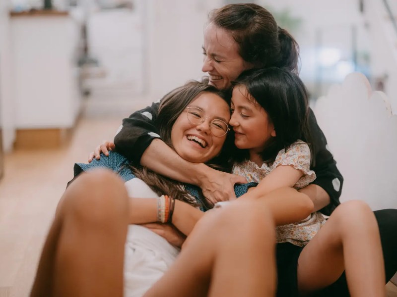 Mutter hält ihre Teenagertochter und eine jüngere Tochter im Arm, alle sitzen auf dem Boden und lachen.